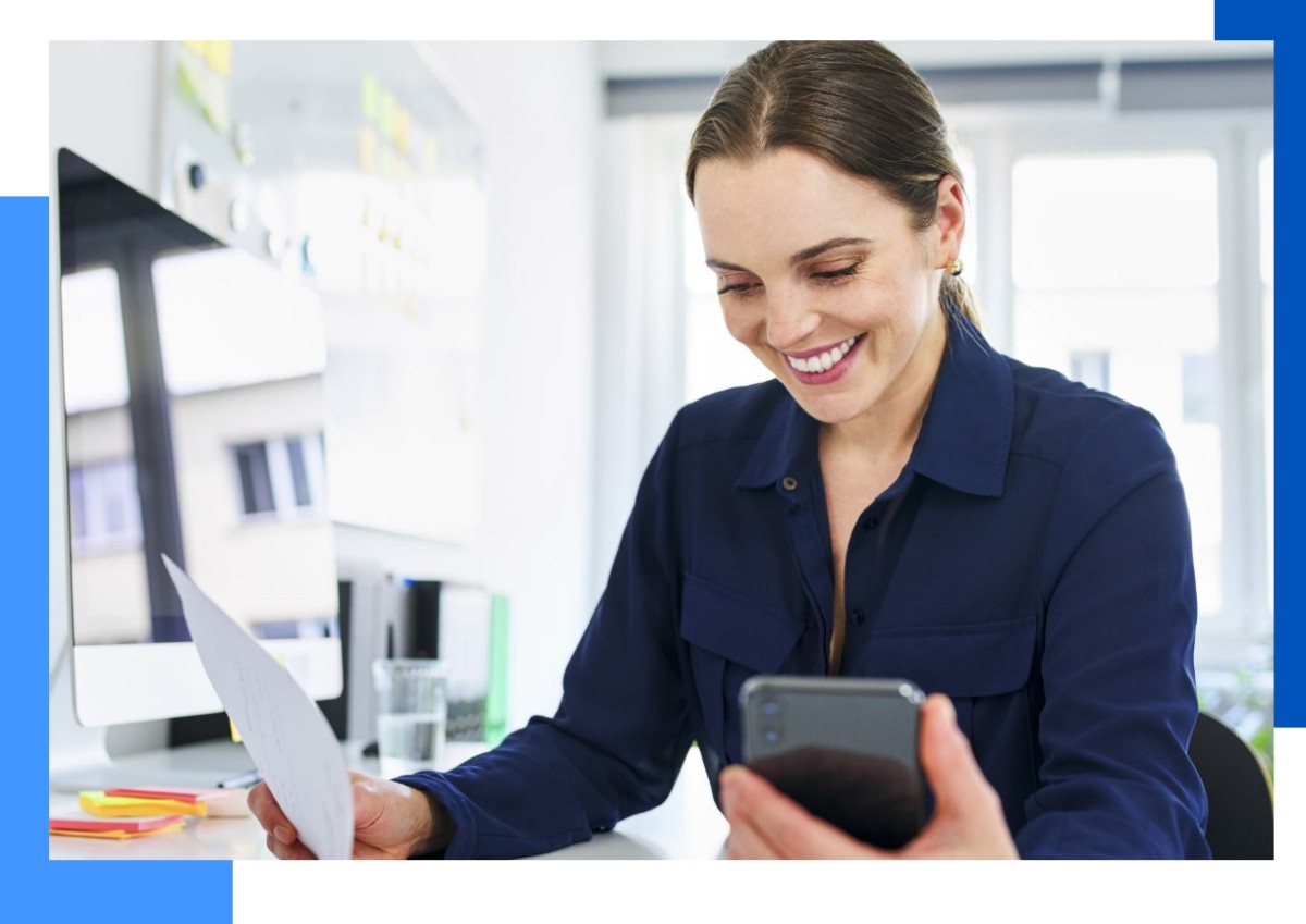 Photo of a woman looking at her phone and working at a laptop