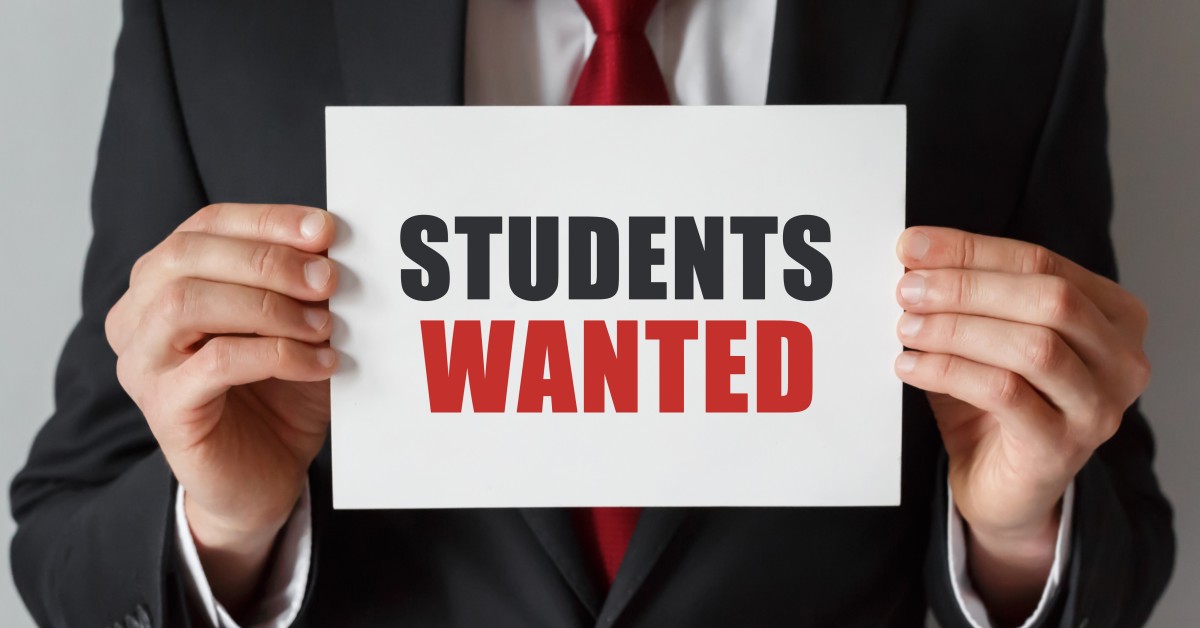 Person in a suit holding a sign that says, Students Wanted