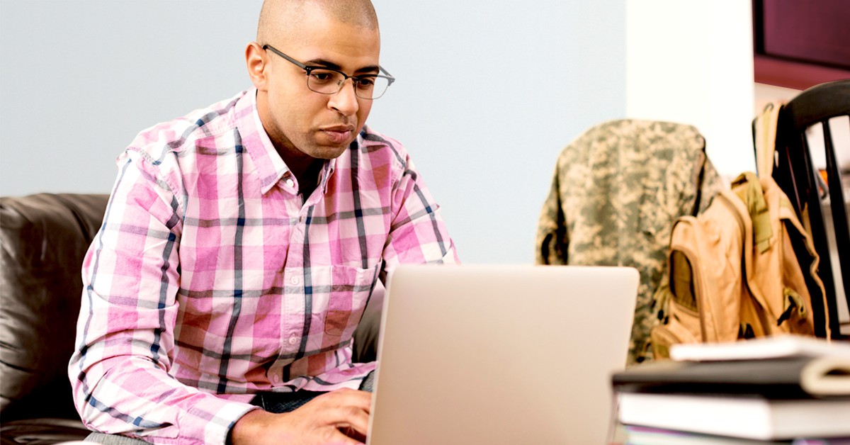 Young man with glasses sitting on a couch typing on a laptop with a military jacket and bags sitting next to him.