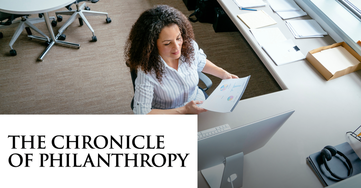 Image of a woman working at a office in front of the computer while reading a physical document. On the lower left corner is placed the Chronicle Of Philanthropy black logo on a white horizontal rectangle. 