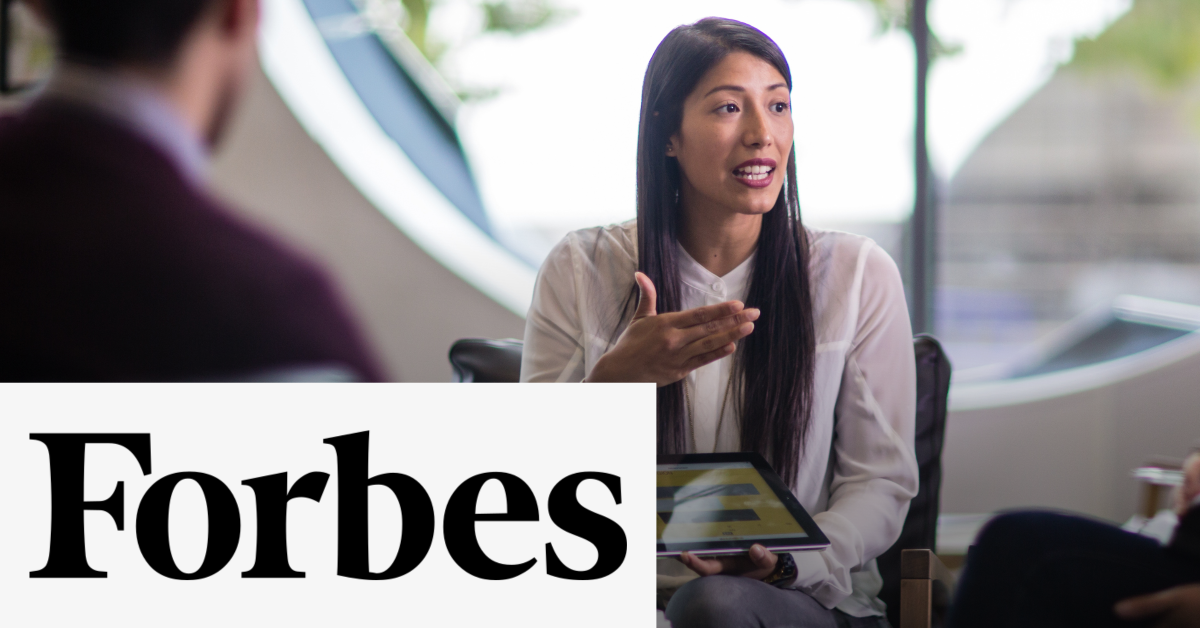 Image of a woman during a speech using an electronic tablet to show her presentation. On the lower left corner the Forbes black logo is placed on a white rectangle. 