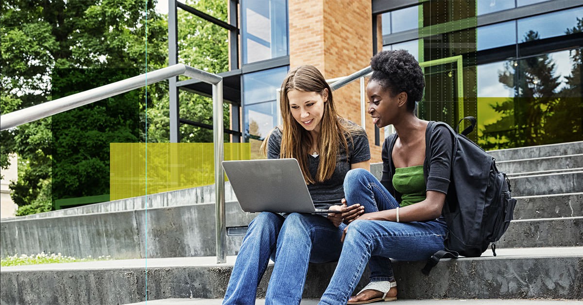 Photo of two students working together at a laptop while sitting on the steps of a building on a college campus