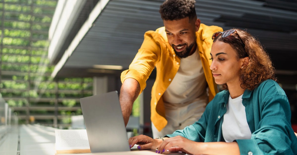 Photo of a person sitting at a laptop with another person over their right shoulder pointing at something on the screen